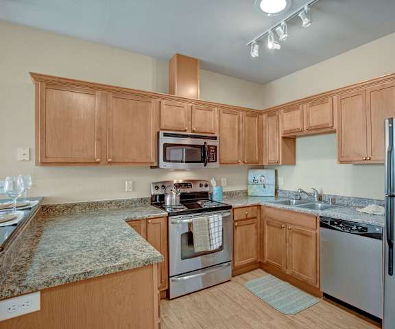 A large kitchen with stainless steel appliances, bar counter with barstools, kitchen utensils, marble countertops with lots of room, and large wooden cabinets at the Vintage Apartment Homes. 