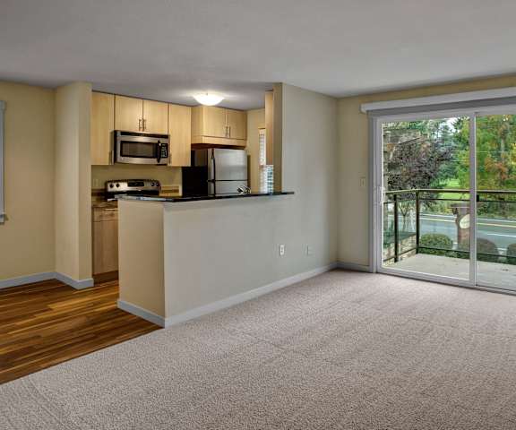 A view of the large, empty carpeted living room with a sliding glass door to the balcony and the kitchen with stainless steel appliances and wooden large cabinets at Vermont Apartment Homes. 