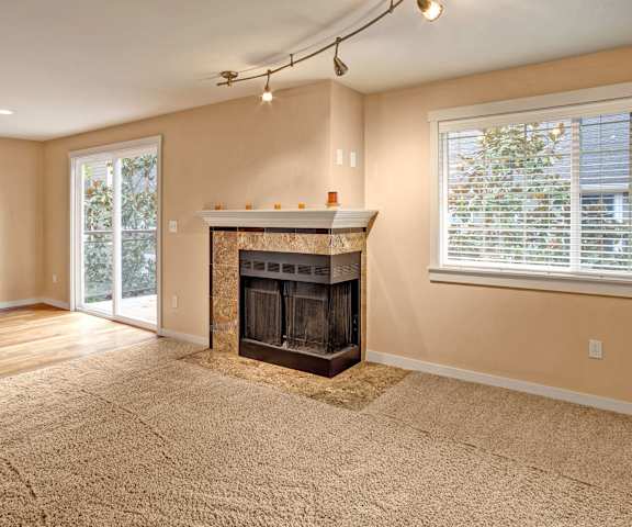 A large, empty living room with carpeting, a window, overhead lighting, a cozy fireplace, and a door to the balcony at Vermont Apartment Homes. 