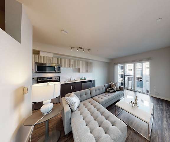 The spacious living room with hardwood floors, a couch, throw pillows, large windows, a lamp, wall art, plush chairs, coffee table, decorations, a view of the open, modern kitchen, and glass doors to the balcony at Vive Apartment Homes. 