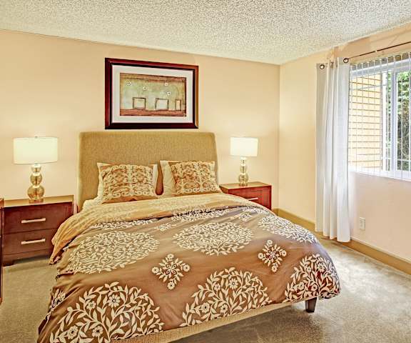 A spacious bedroom with a queen-sized bed with a golden bedspread, a dresser, wall art, lamps, decorations, window, and nightstands at Whispering Cedars Apartment Homes. 