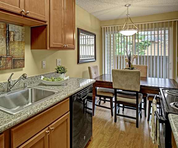 A spacious kitchen with black appliances, marble countertops, wall art, a fruit bowl, and wooden cabinets with decorations, a dining room with a dining table and chairs, and the doors to the balcony at Whispering Cedars Apartment Homes. 