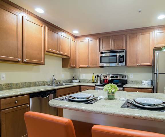 A large kitchen with stainless steel appliances, marble countertops, large wooden cabinets, and two places set at Windsong Apartment Homes. 