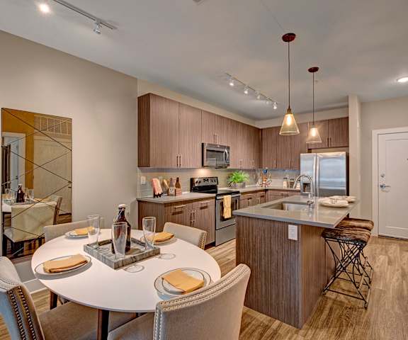 A large kitchen with stainless steel appliances, marble countertops, large wooden cabinets, two barstools, two places set at the kitchen island, and the dining room, with a mirror on the wall, four chairs, and four places set at the table at River House Apartment Homes. 