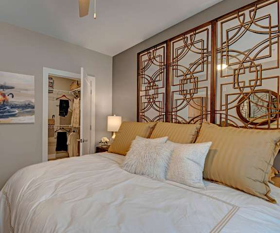 A spacious bedroom with a king-size white bed, a large walk-in closet, wall art, lamps, decorations, window, and nightstands at River House Apartment Homes. 