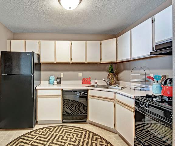A cozy kitchen with black appliances, white countertops with lots of room, and white wooden cabinets with a Welcome Home Sign, a striped rug, kitchen appliances, decorations at River's Cove Apartment Homes. 