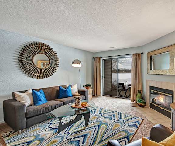The spacious living room with a gray couch, blue and white throw pillows, a large window, a ceiling fan, wall art, plush chairs, coffee table, decorations, a blue, white, and yellow striped rug, a TV, and glass doors with curtains to the balcony at River's Cove Apartment Homes. 