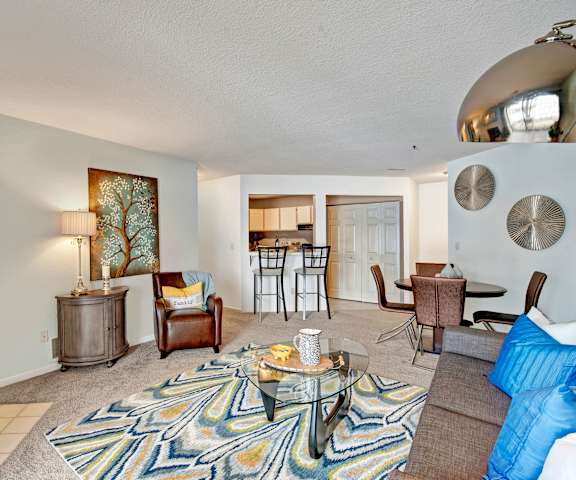 The spacious living room with a gray couch, brown leather chair, coffee table, overhead lighting, decorations, a TV, and view of the dining room with a table and four chairs and a bar with two barstools at River's Cove Apartment Homes. 