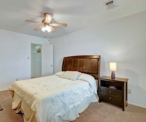 A spacious bedroom with a queen-size bed with a white bedspread, a dresser, wall art, decorations, overhead lighting with a ceiling fan, closet, and nightstands at St. James Place Apartment Homes. 
