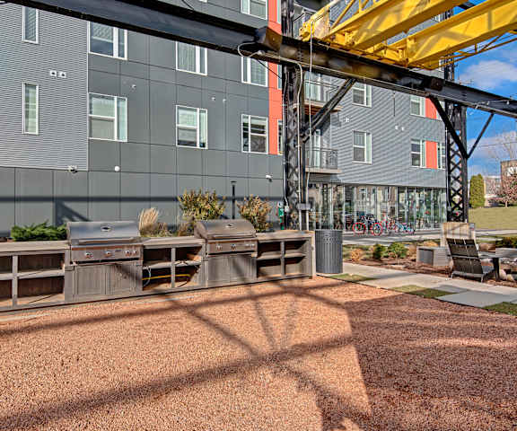 The resident grilling area underneath a metal structure in front of the dark gray and orange apartment building at Stitchweld Apartment Homes. 