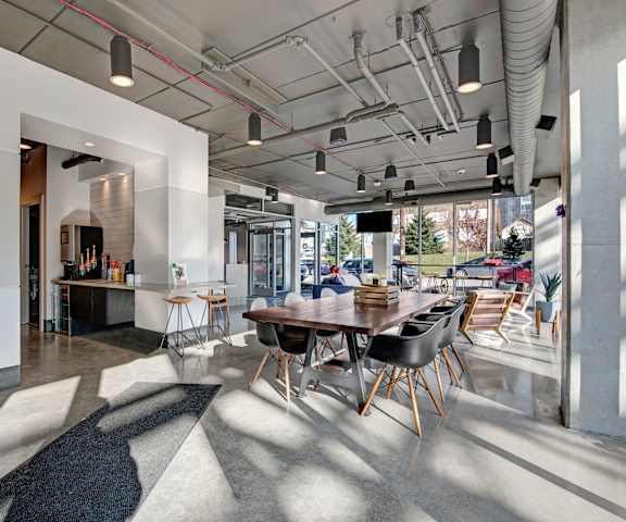 A large, gray, industrial resident lounge with overhead lighting, a long table with lots of chairs, large windows, and a coffee station at Stitchweld Apartment Homes. 