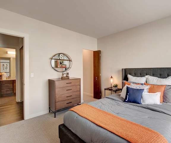 A spacious bedroom with a queen-size bed with a gray, orange, and white bedspread, a brown dresser, wall art, decorations, closet, and nightstands at Stitchweld Apartment Homes. 
