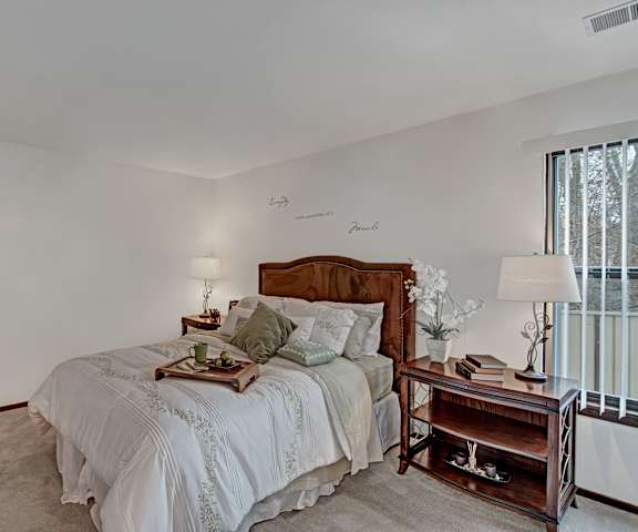 A spacious bedroom with a queen-size bed with a light gray bedspread, a dresser, wall art, lamps, decorations, window, and nightstands with lamps at Sunset Ridge Apartment Homes.