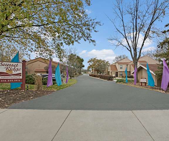 The entrance to Sunset Ridge Apartments with two red and white signs surrounded by trees alongside a road with the apartments and purple and blue flanking it at Sunset Ridge Apartments. 