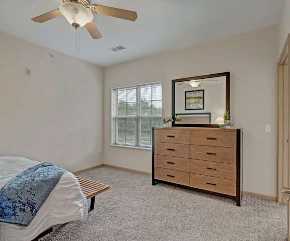 A spacious bedroom with a queen-size bed with a bedspread, a dresser, overhear lighting with a fan, wall art, lamps, decorations, and window at Swan Creek Apartment Homes. 