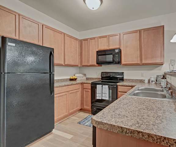 A spacious kitchen with black appliances, overhead lighting, granite countertops with lots of room, and wooden cabinets with two places set at the bar at Swan Creek Apartment Homes. 