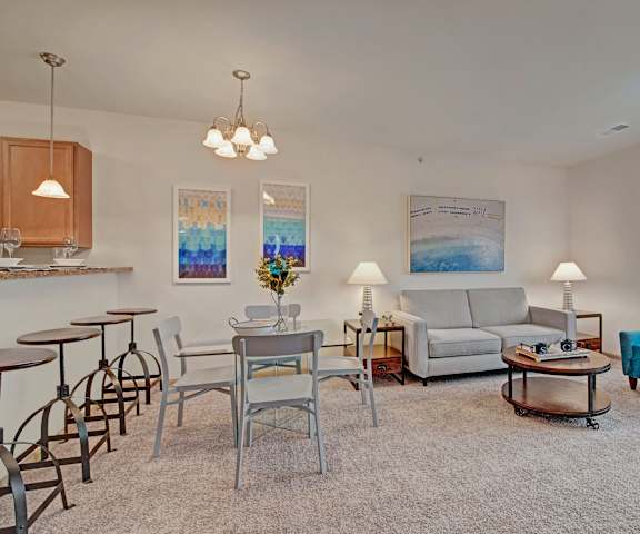 The spacious living room with a gray couch, a blue plush chair, coffee table, overhead lighting, decorations, and view of the dining room with a gray table, chairs, and place settings for four, and the kitchen at Swan Creek Apartment Homes. 