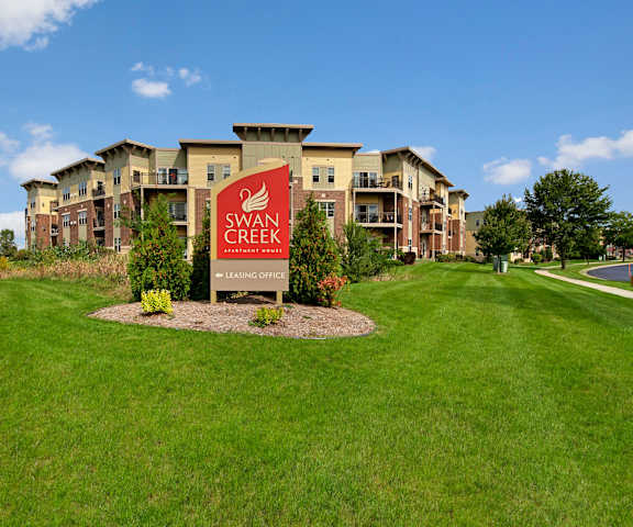 The red main sign in front of the apartments surrounded by green grass, shrubs, and trees at Swan Creek Apartment Homes. 