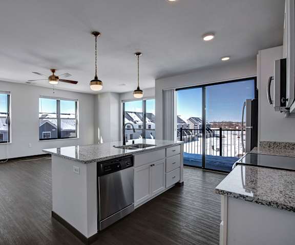 A large, modern kitchen with stainless steel appliances, kitchen utensils, granite countertops with lots of room, a kitchen island, and large white wooden cabinets and an empty hardwood living room with large windows at the Lafayette Apartment Homes. 
