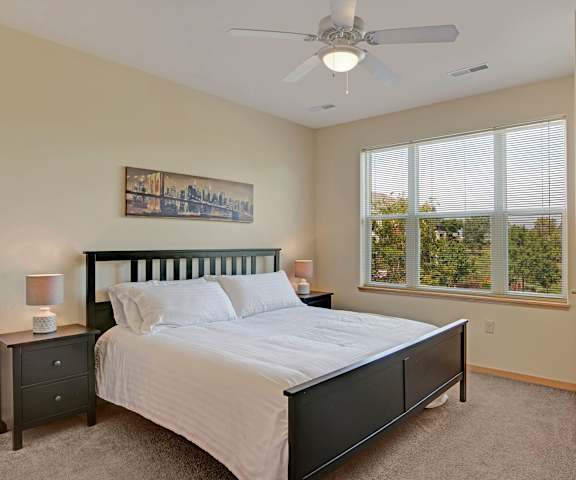 A spacious bedroom with a queen-size bed with a white bedspread, a dresser, wall art, lamps, decorations, window, and nightstands at Van Buren Place Apartment Homes. 