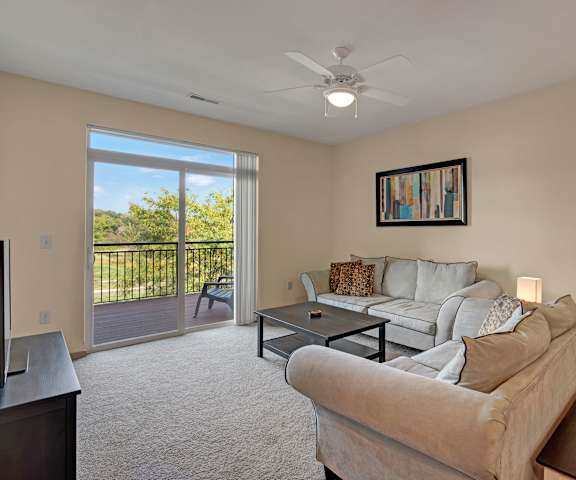 The spacious, white, modern, living room with two gray couches, throw pillows, large windows, a ceiling fan, wall art, plush chairs, coffee table, decorations, a TV, and glass doors to the balcony at Van Buren Place Apartment Homes. 