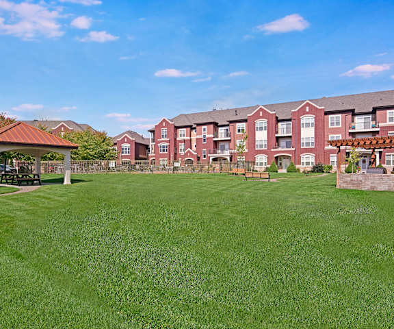 An outdoor resident patio with a grill area, green space with grass and plants, and tables and chairs with overhead string lights surrounded by the white and orange apartment buildings at Van Buren Place Apartment Homes. 