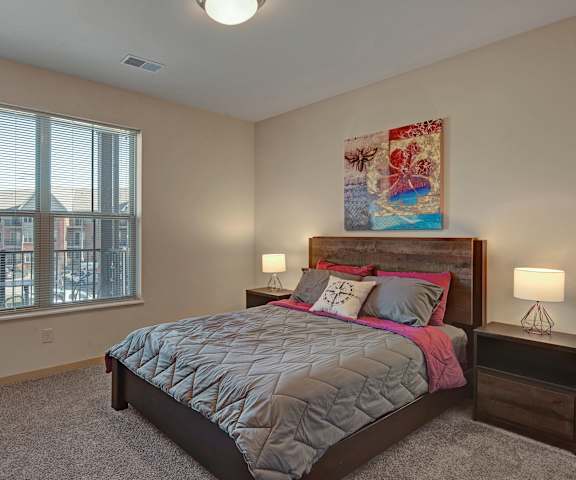 A spacious bedroom with a queen-size bed with a gray and pink bedspread, a dresser, wall art, lamps, decorations, window, and nightstands at The Vue at Pinnacle Park Apartment Homes. 