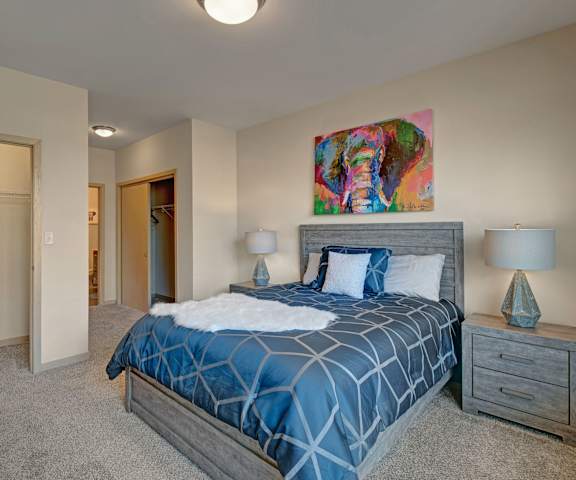 A spacious bedroom with a queen-size bed with a blue bedspread, a dresser, wall art, lamps, decorations, closets, and nightstands at The Vue at Pinnacle Park Apartment Homes. 