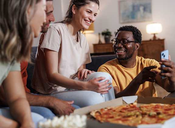 A group of people are sitting on a couch, eating pizza and playing a card game.