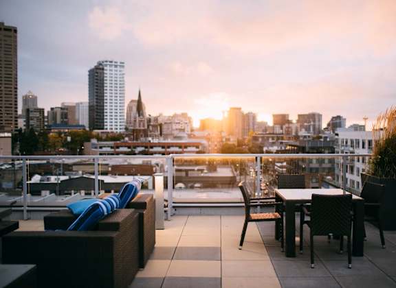 A rooftop patio with a table and chairs overlooking a city skyline at sunset.