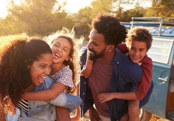 A family of four is hugging and smiling together in front of a blue vehicle.