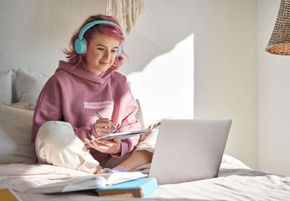 A person with pink hair is wearing headphones and writing in a notebook while using a laptop.at OSM Properties Homes & Apartments, Chico, CA