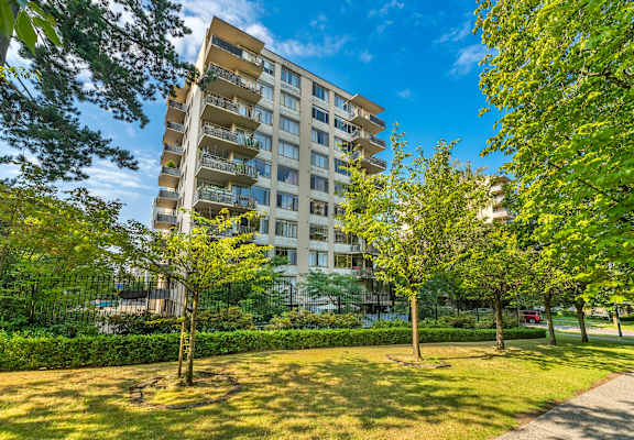 A tall apartment building with balconies is surrounded by green trees and bushes.