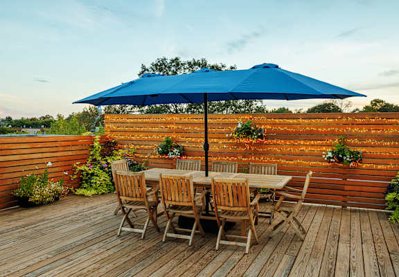 A blue umbrella is on a wooden deck with chairs and a table.