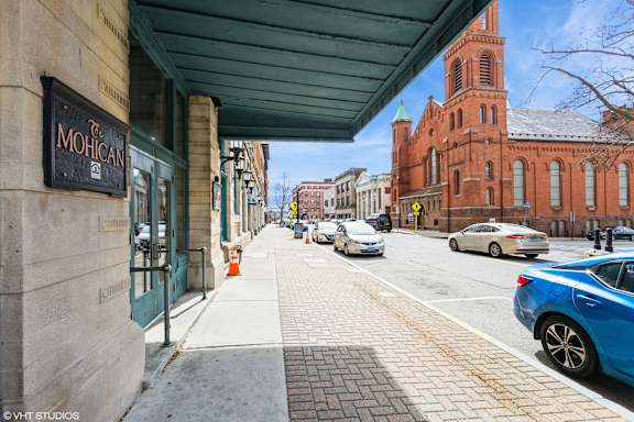A street view with cars parked on the side and a building with a sign that reads at Mohican Apartments, New London, 06320