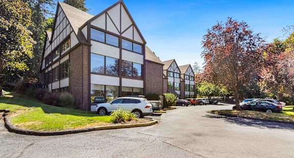 A modern building with a mix of brick and glass windows is surrounded by trees and has a parking lot in front.
