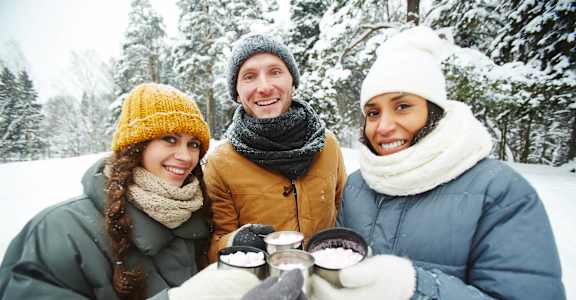 Three people in winter clothing holding cups in a snowy forest at The Woods at Copper Ranch, Hailey