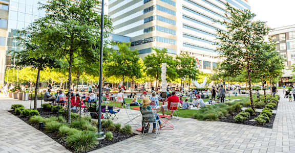 A park with people sitting and standing on the grass and pavement.