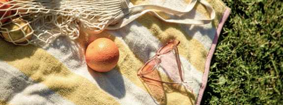 A pair of sunglasses and an orange are on a blanket next to a mesh bag.