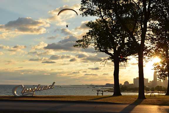 A person is paragliding over a park with trees and a Cleveland sign in the background.