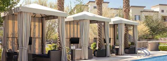 Four palm trees are behind a pool with four sun shelters.