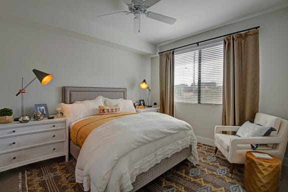 A spacious bedroom with a queen-size bed with a white and yellow bedspread, a dresser, wall art, lamps, decorations, window with curtains, a white plush chair in the corner, and nightstands with lamps at Roosevelt Row Apartment Homes. 