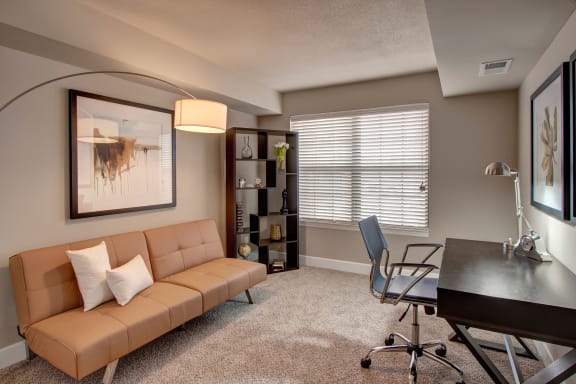 A modern office with a desk on a lamp, chair, tan couch with white pillows, windows, shelves, coffee table, wall art, decorations, and large window with light streaming in at Fairways Apartment Homes. 