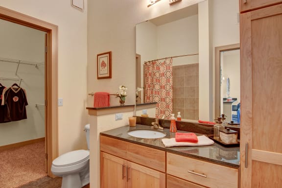 The cozy bathroom with a sink, mirror, shiny black countertop with toiletries, a shower with an orange patterned shower curtain, and a toilet at Residence at the COR Apartment Homes. 
