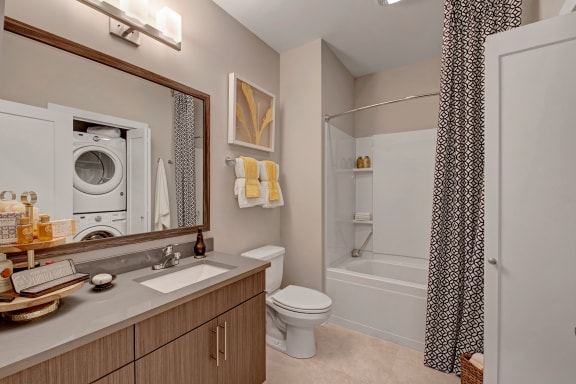 The luxury bathroom with lots of counter space, a shower with a black and white patterned shower curtain, a large mirror, a sink, a toilet, and a closet with a washer and dryer at River House Apartment Homes. 