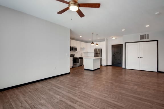 A view of the empty hardwood living room with overhead lighting a ceiling fan and the large kitchen with stainless steel appliances, marble countertops, and spacious wooden cabinets at the Lafayette Apartment Homes. 