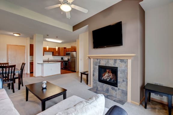 The spacious living room with a couch, fireplace, plush chairs, coffee table, overhead lighting with a ceiling fan, decorations, a TV, and view of the dining room with a table, chairs, and place settings for four, and the kitchen at Van Buren Place Apartment Homes. 
