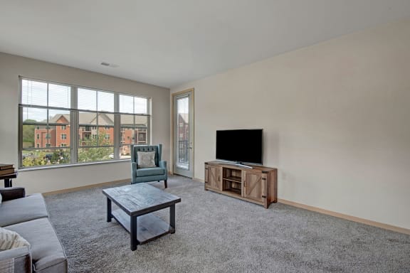 The spacious living room with a couch, throw pillows, large window s, a ceiling fan, wall art, plush chairs, coffee table, decorations, large windows, a TV, and glass doors to the balcony at The Vue at Pinnacle Park Apartment Homes. 