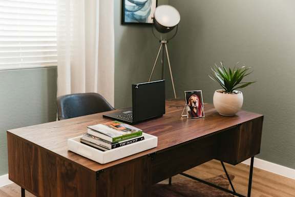 A black laptop sits on a wooden desk with a stack of books and a small potted plant.