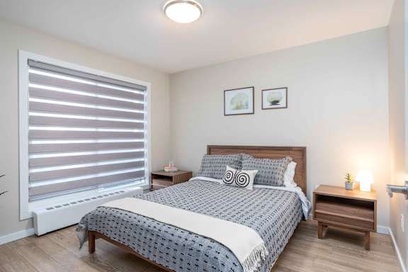 A bedroom with a bed, nightstand, and window with blinds at G17 Apartments at Tamarack, Edmonton, AB